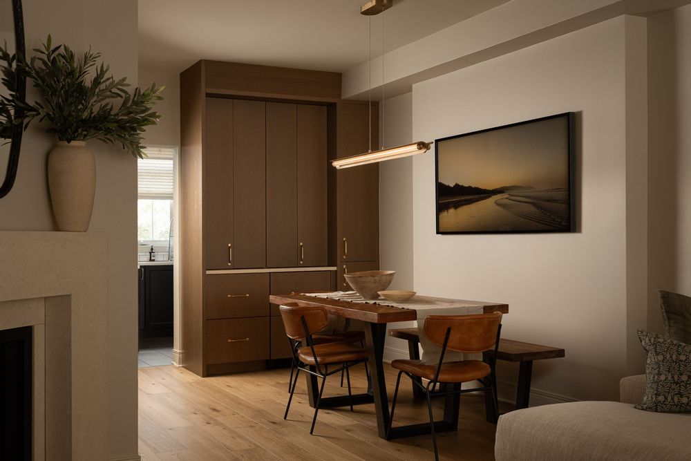 Modern dining area with wooden table, black chairs, warm pendant light, built-in cabinetry, and coastal artwork on wall.