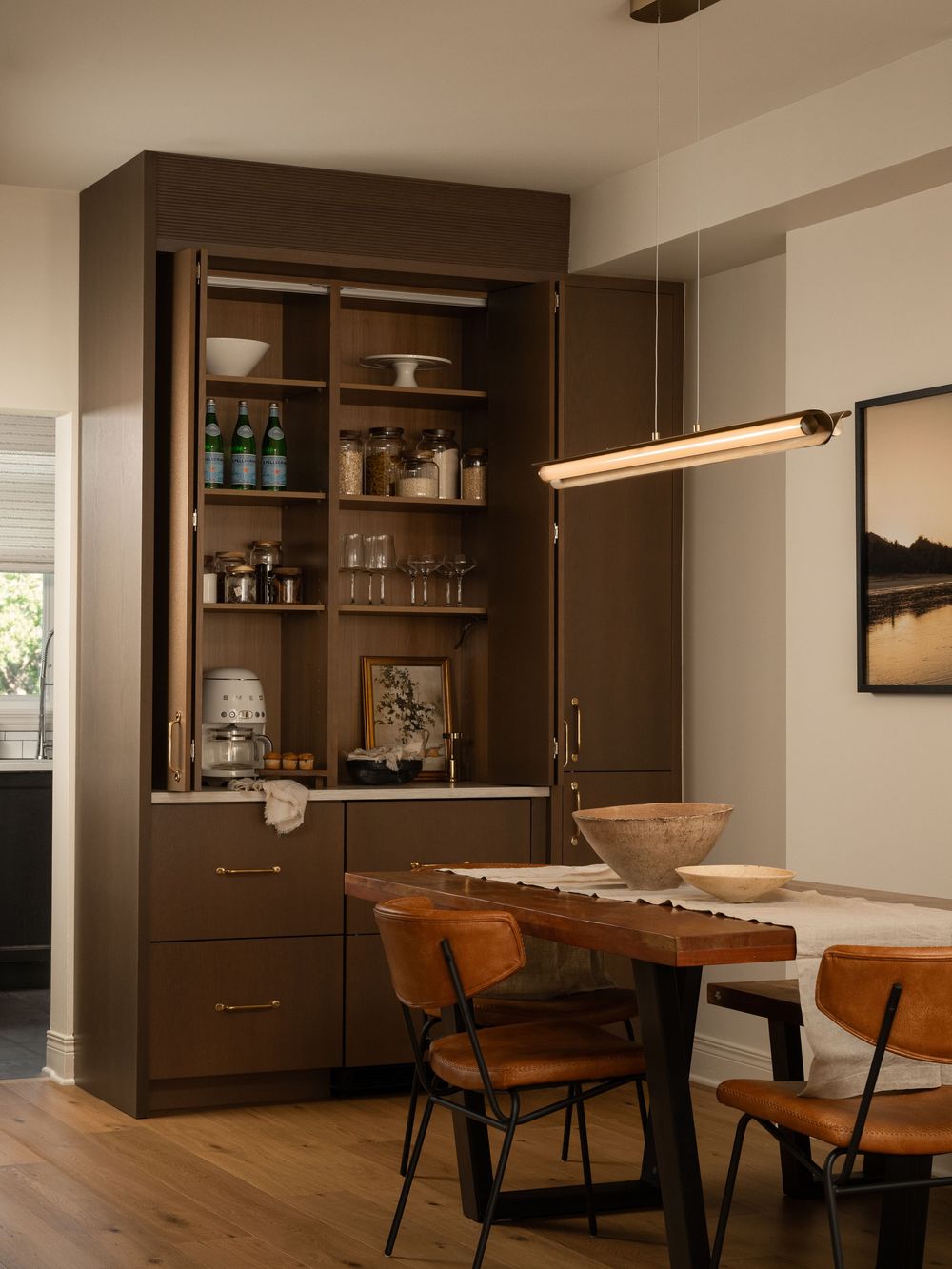 Dark brown built-in cabinet with open shelves displaying bottles and glassware beside a wooden dining table with leather chairs.