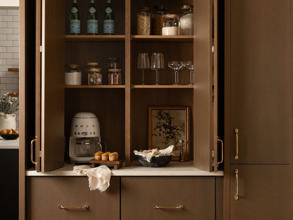 Open dark brown kitchen cabinet displaying organized pantry items, glassware, a coffee maker, and gold hardware accents.