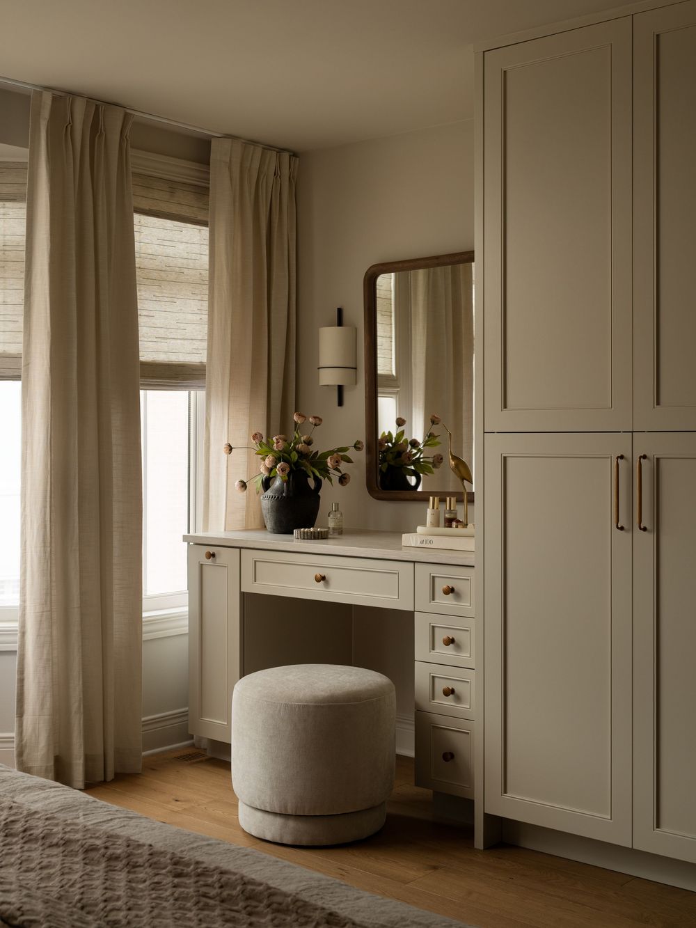 Elegant bedroom vanity with sage green cabinetry, round mirror, linen curtains, and upholstered ottoman on hardwood floors.