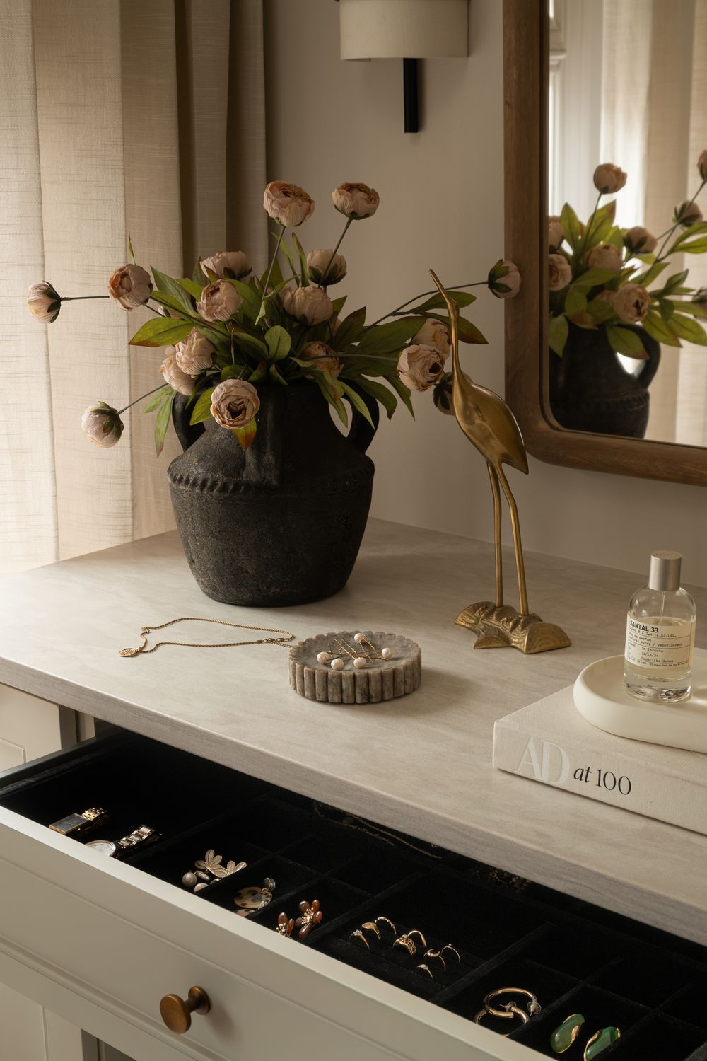 Elegant vanity dresser with open jewelry drawer, brass heron figurine, dark vase with roses, and perfume bottle on marble top.