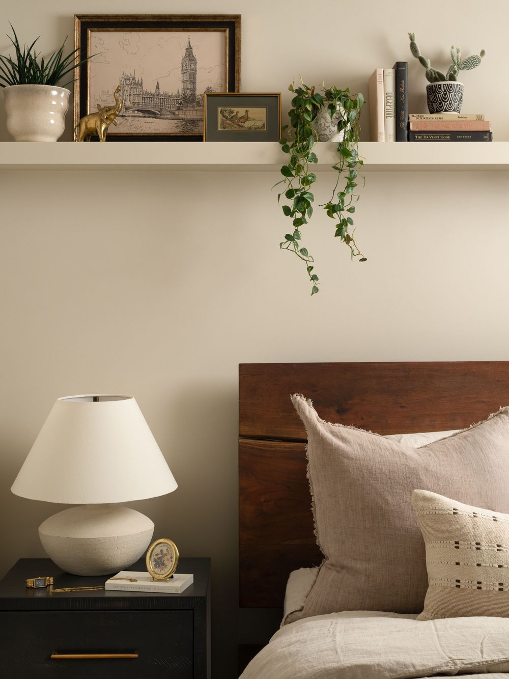 Cozy bedroom with dark wood headboard, white lamp on nightstand, floating shelf with plants and art above the bed.
