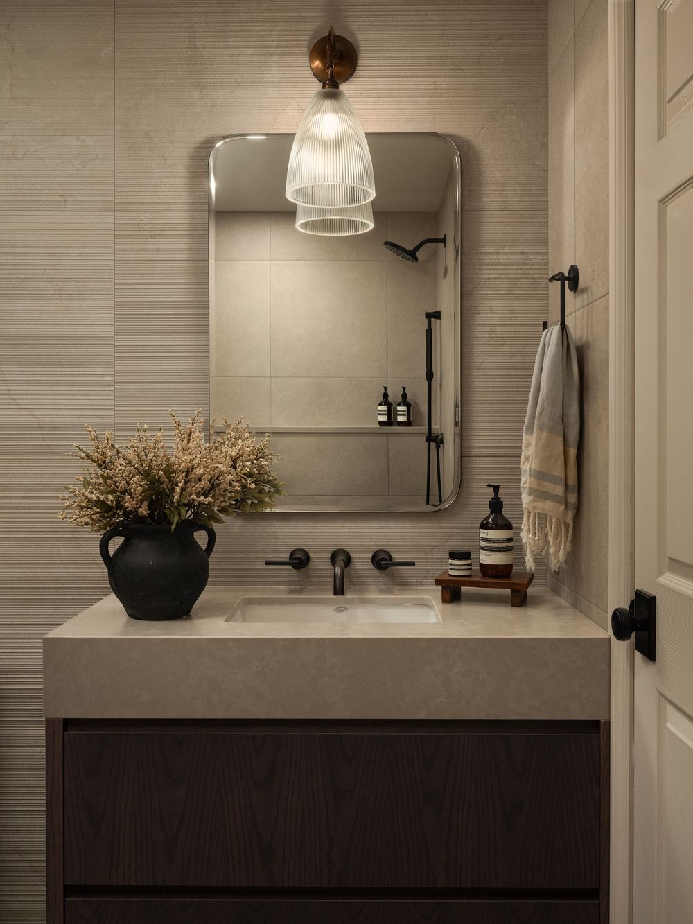 Modern bathroom vanity with beige stone countertop, dark wood cabinet, matte black faucet, round mirror, and glass pendant light.
