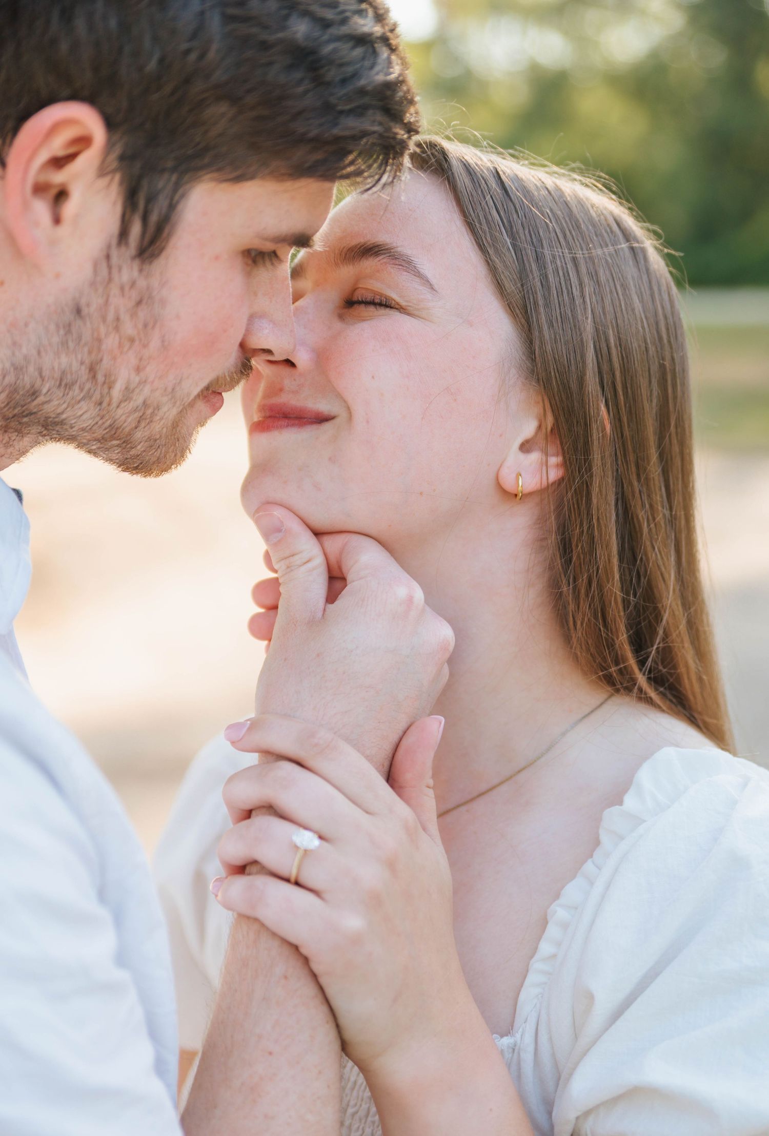 A couple shares a tender romantic moment, the man gently holding the woman's chin as they lean in for a kiss outdoors.