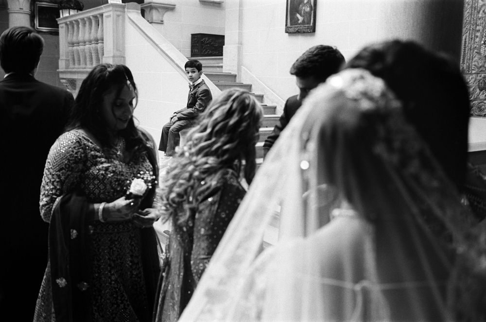 Black and white wedding photo of bride in veil with guests in traditional attire on staircase.