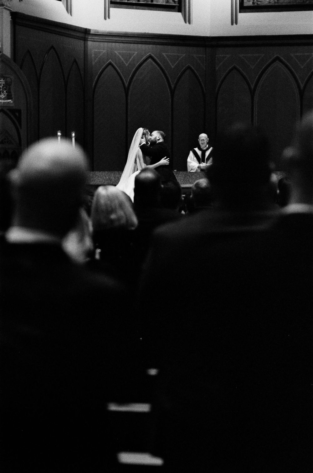 Black and white photo of bride and groom sharing first kiss at church altar with guests watching.