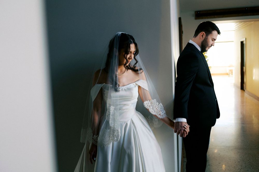Bride in off-shoulder white gown with veil holds groom's hand in hallway on wedding day.