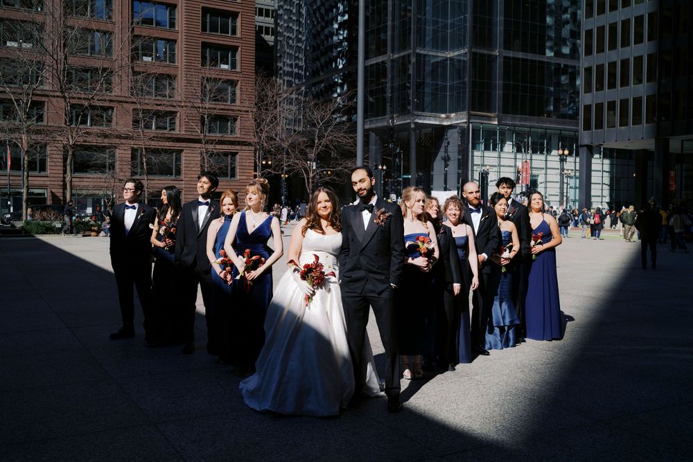 Wedding party poses outdoors in urban setting, bride in white gown with groomsmen in tuxedos and bridesmaids in navy dresses.