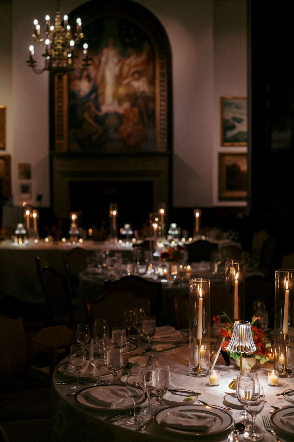 Elegant candlelit wedding reception tables with tall candles, fine glassware, and classic artwork in a dimly lit venue.