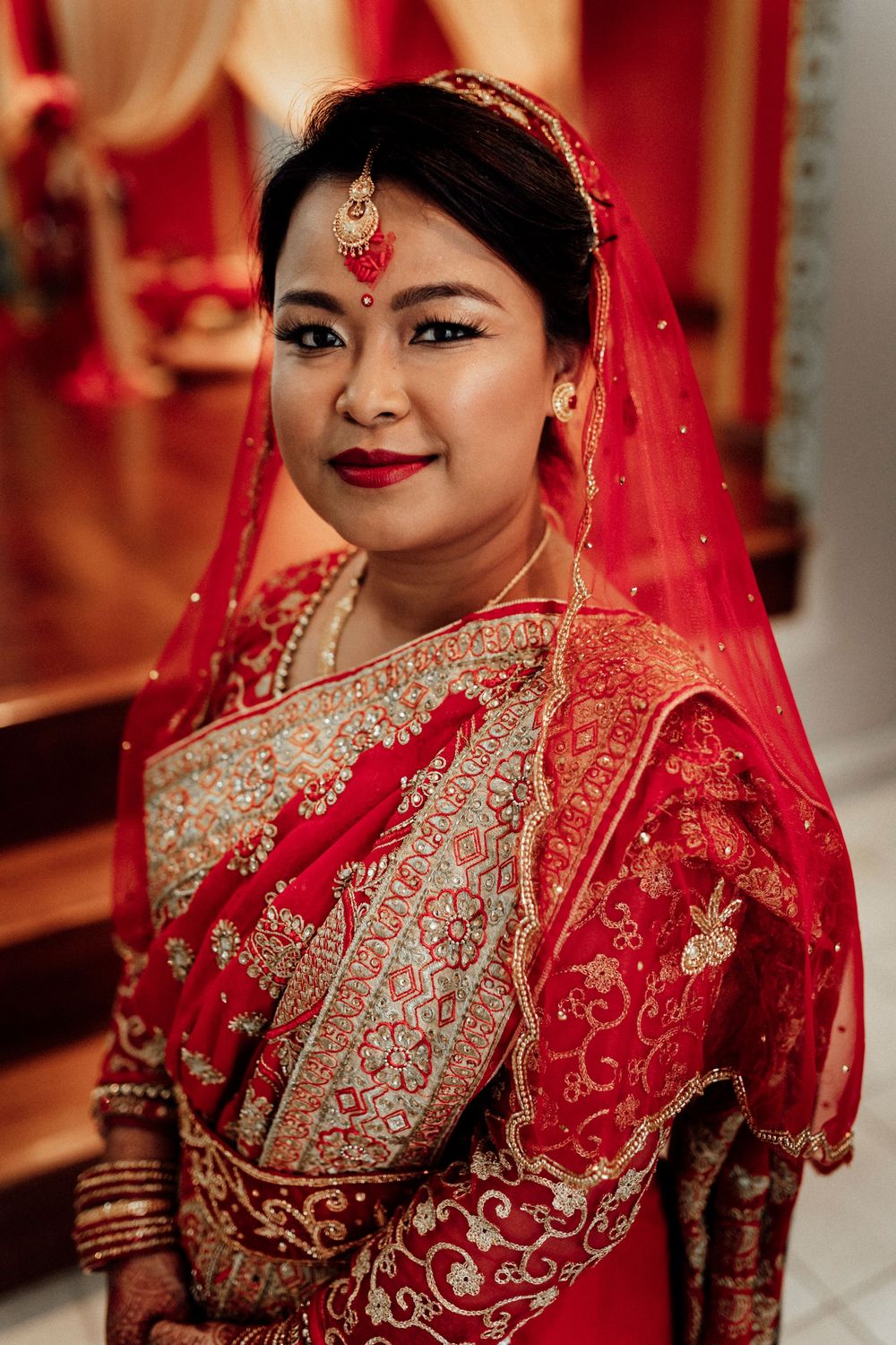 Indian bride in ornate red and gold saree with traditional jewelry and red veil smiles at camera.
