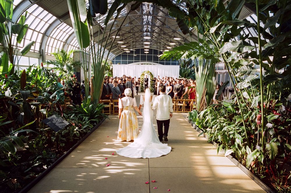 Bride and groom walk down aisle in lush greenhouse wedding venue filled with tropical plants and seated guests.