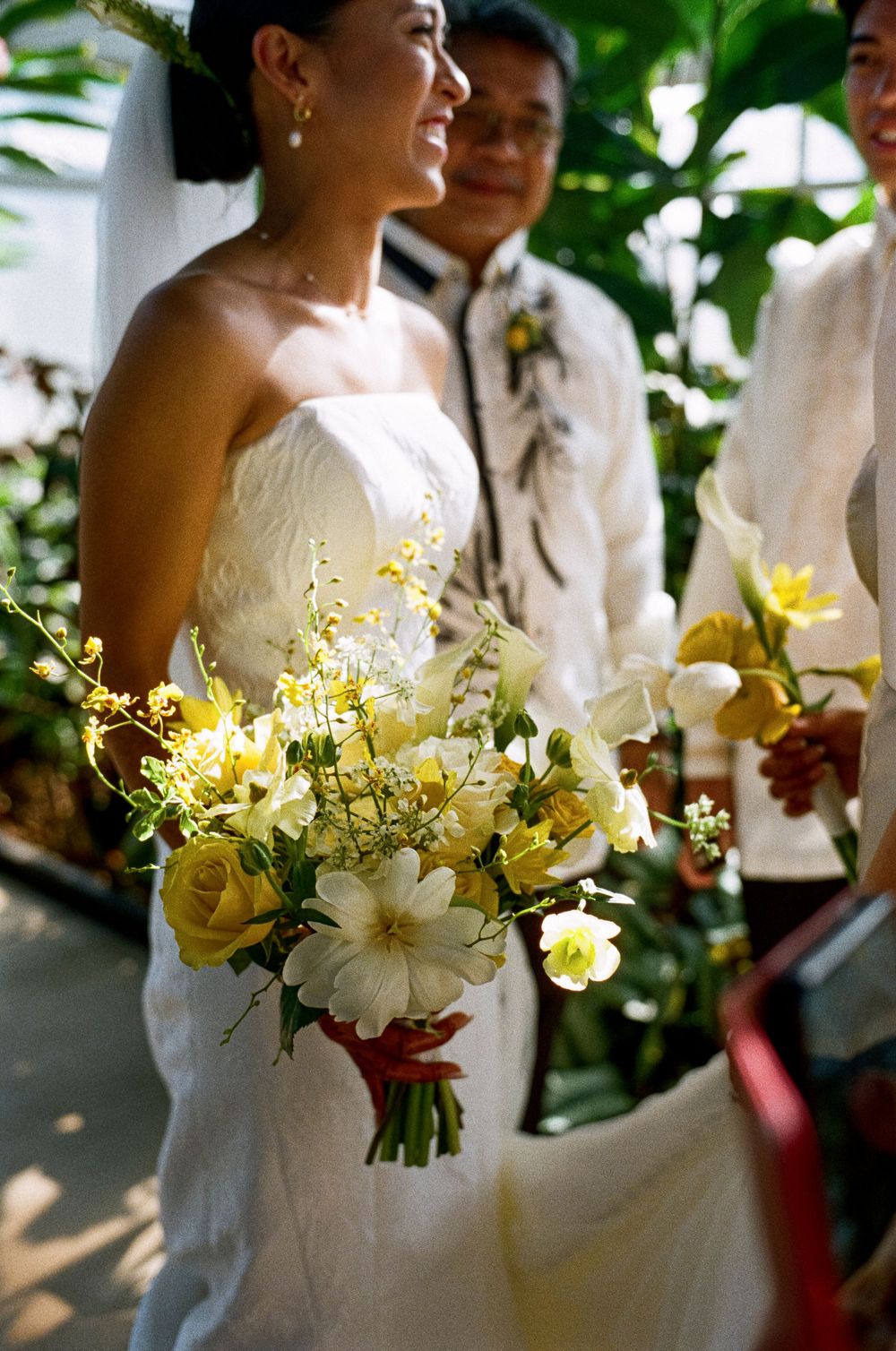 Smiling bride holding a yellow and white floral bouquet with roses and daisies during outdoor wedding ceremony.