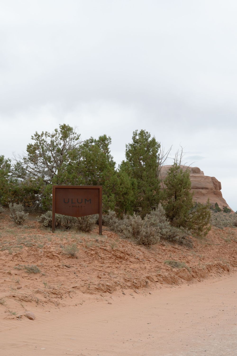 Brown wooden sign at Uluru desert trail surrounded by green shrubs, red sandstone rocks, and sandy terrain under cloudy skies.