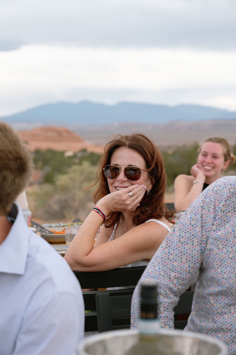 A smiling woman wearing sunglasses enjoys an outdoor dining experience with scenic mountain views in the background.
