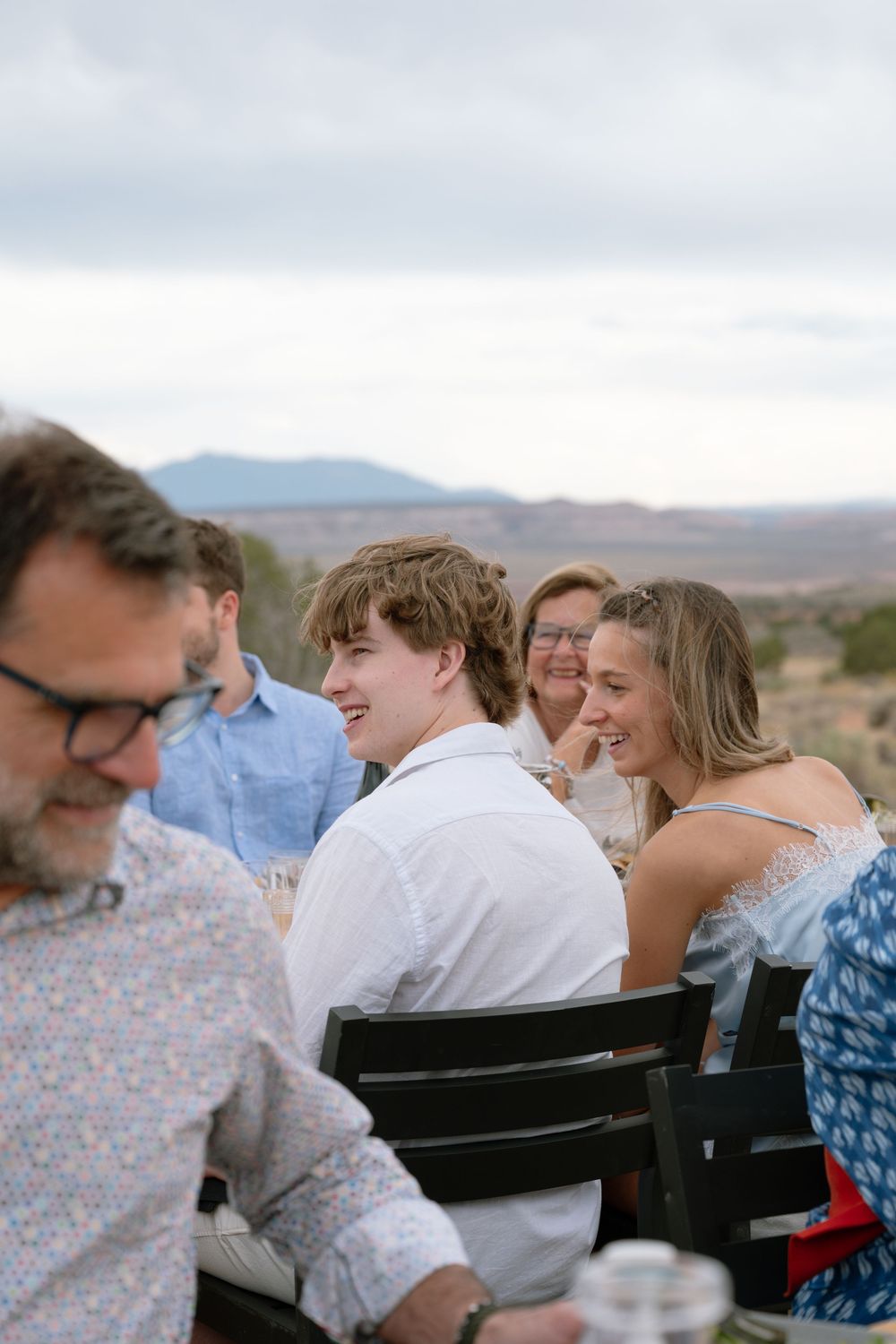 Group of young adults laughing and socializing outdoors with scenic desert mountain landscape in background.
