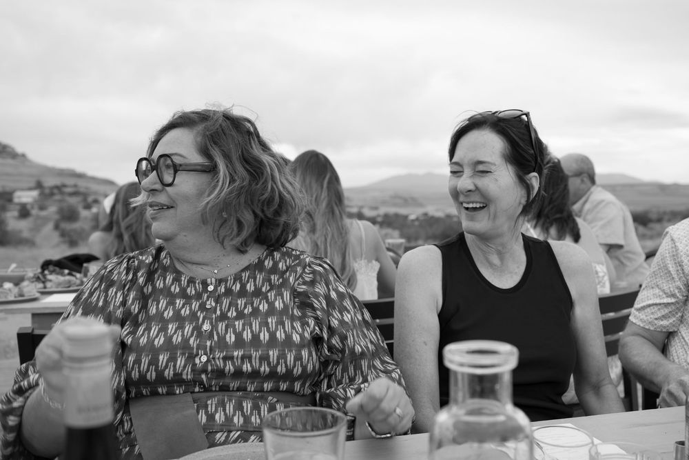 Two women laughing together at an outdoor dining table with scenic hills in the background, black and white photo.