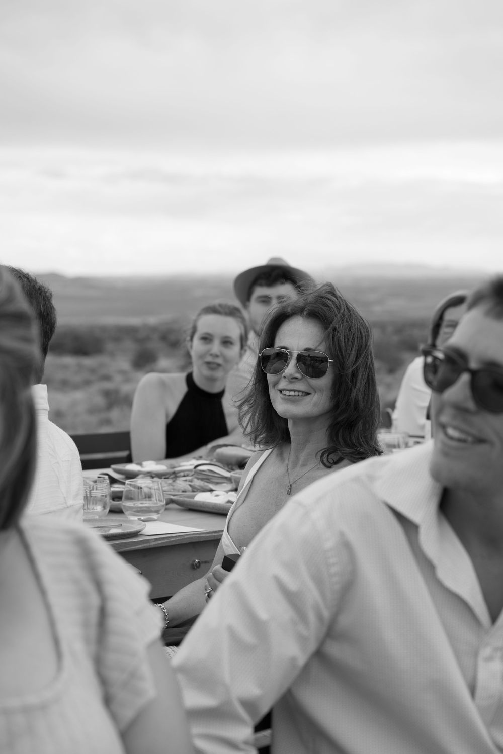 Black and white photo of smiling woman in sunglasses at outdoor dining table with friends and scenic landscape backdrop.