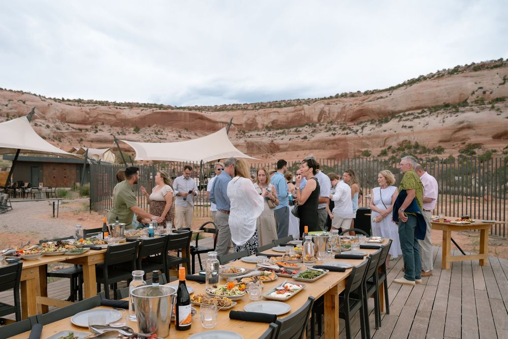 Outdoor desert dining event with guests mingling around long wooden tables set with food and drinks.