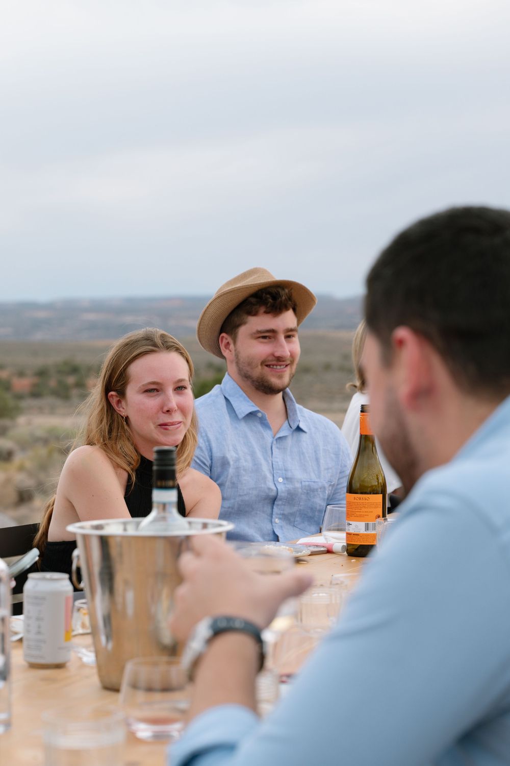 Friends dining outdoors at a rustic table with wine bottles, enjoying a scenic desert landscape view.