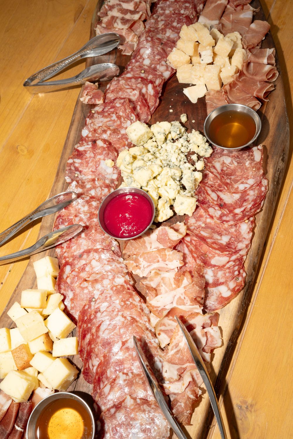 A rustic wooden charcuterie board with assorted meats, cheeses, and dipping sauces served with tongs.