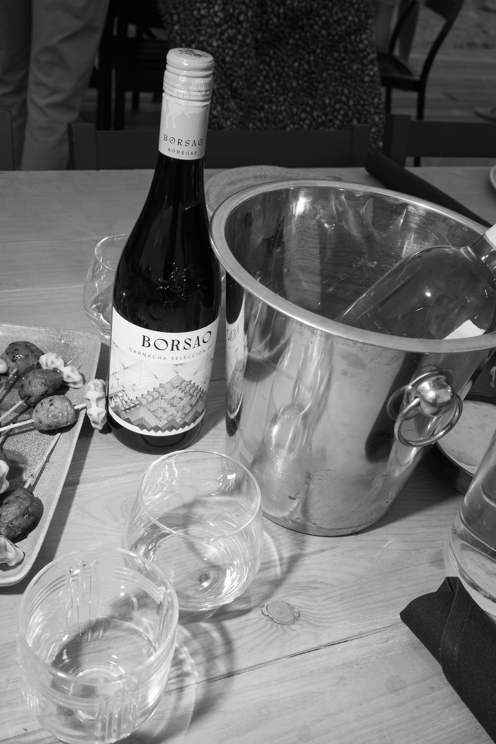 Borsao wine bottle beside silver ice bucket with empty glasses on wooden table, black and white photo.