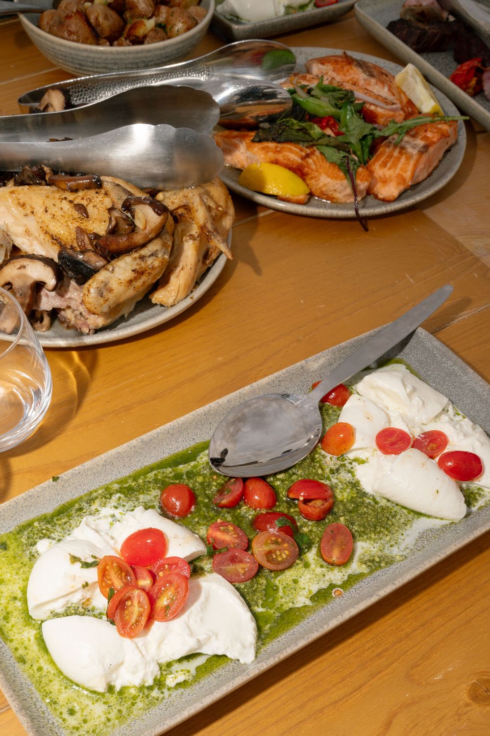 Caprese salad with fresh mozzarella, cherry tomatoes, and pesto on a rectangular plate, with grilled fish and mushrooms in background.