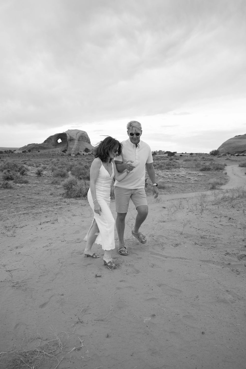 Black and white photo of a couple walking together on sandy desert terrain with rock formations in the background.