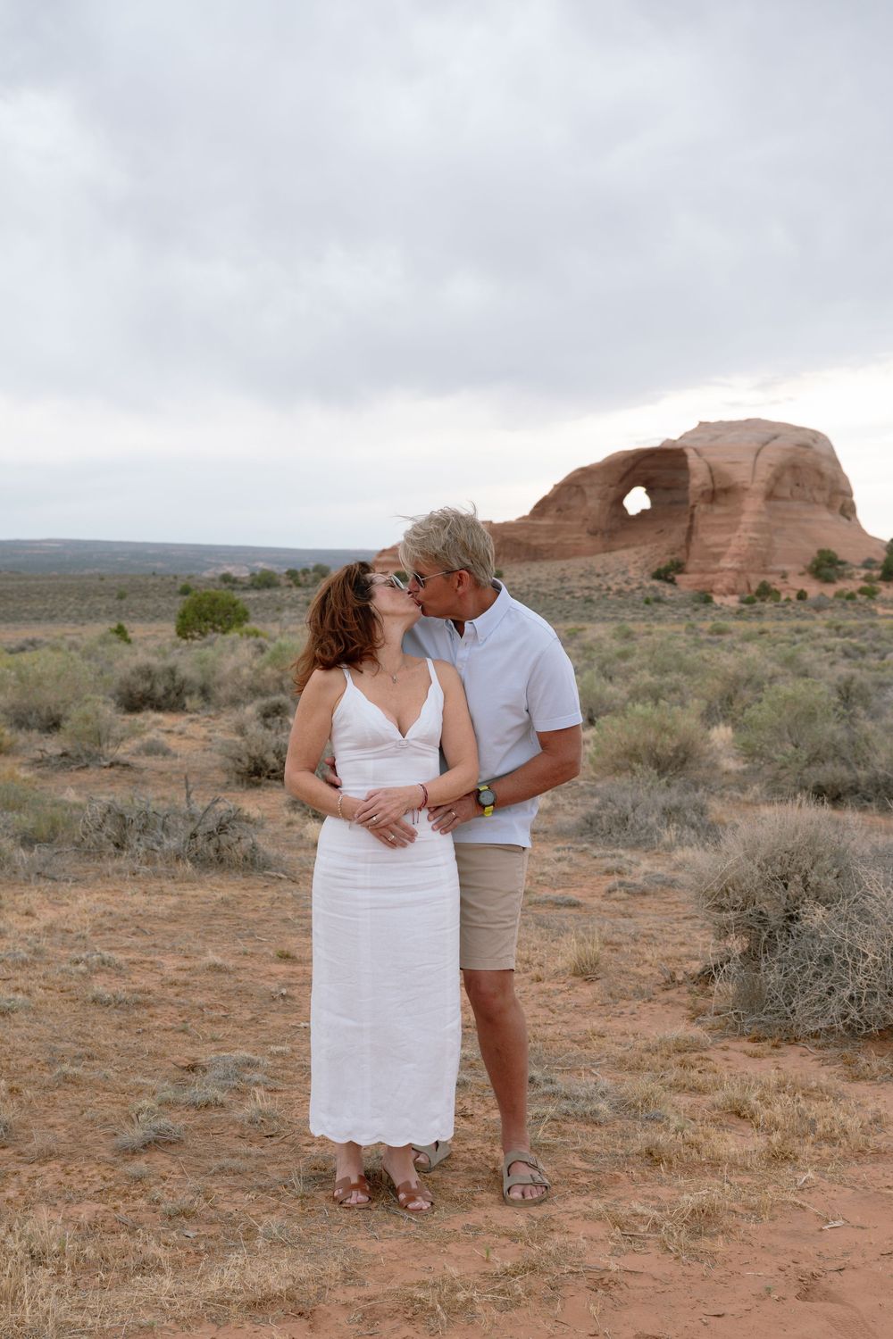 Couple kissing in a desert landscape with a natural rock arch formation in the background.