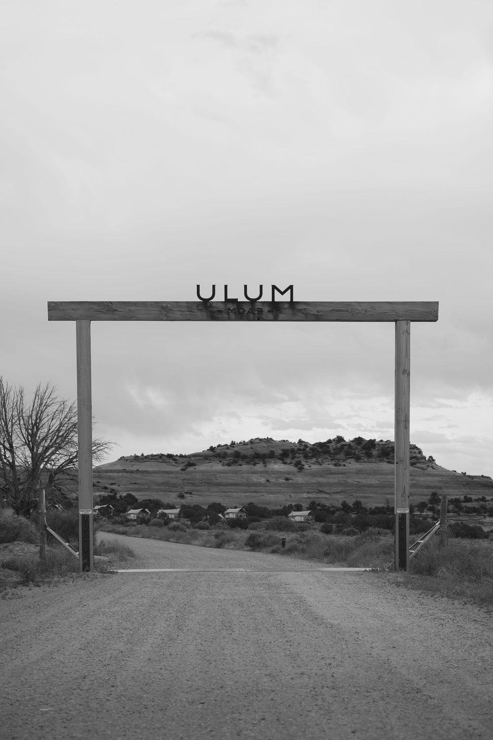 A black and white photo of a wooden entrance gate reading ULUM along a gravel road in a desert landscape.