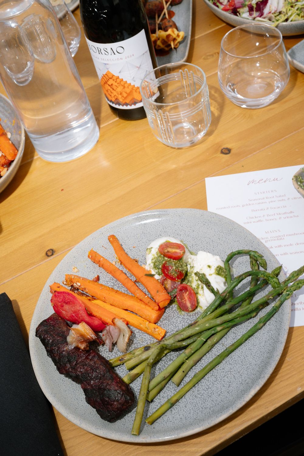 A gourmet plate with roasted carrots, grilled meat, asparagus, and burrata, served alongside a bottle of Orsao wine.