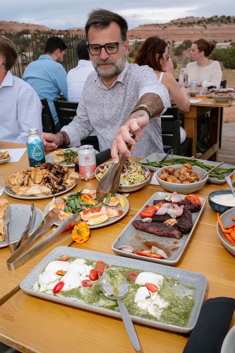 Man serving food at an outdoor dining table filled with various dishes including grilled meats and salads.