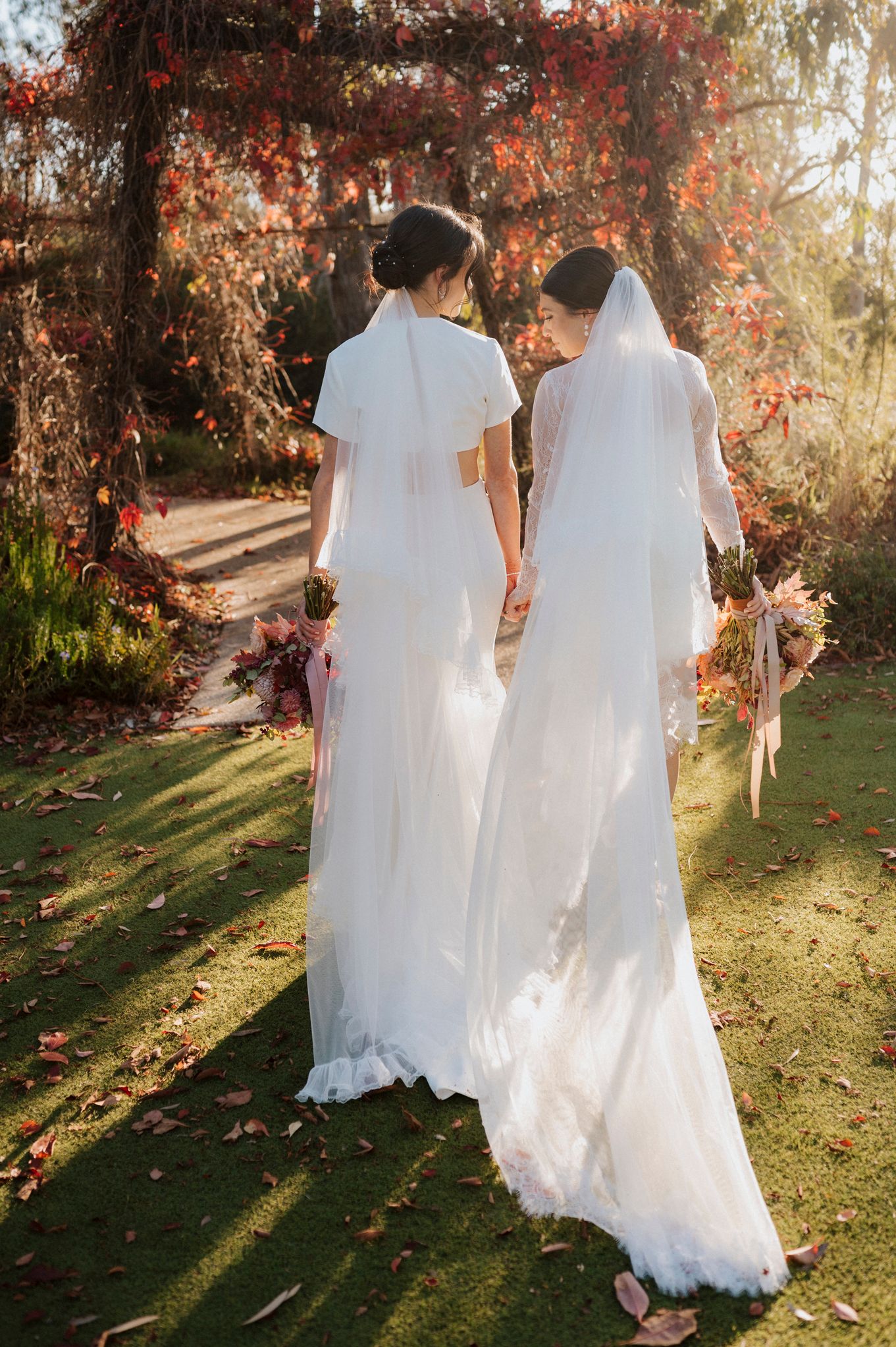 Two brides in white gowns and veils holding bouquets stand before a red floral arch in a sunlit garden.