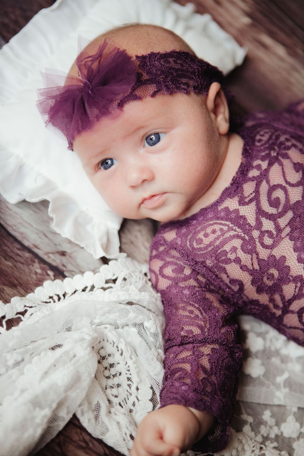Newborn baby girl in purple lace outfit and floral headband lying on white ruffled pillow.