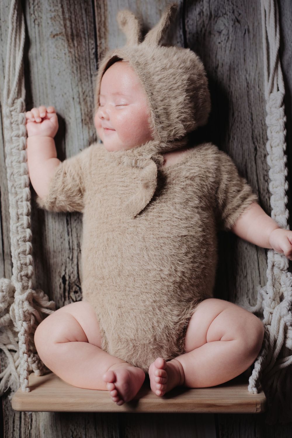 Sleeping newborn baby in fuzzy brown bunny costume on macrame swing against rustic wood.