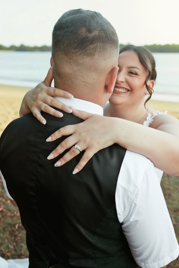 Smiling bride hugging groom from behind on beach, showing diamond engagement ring.