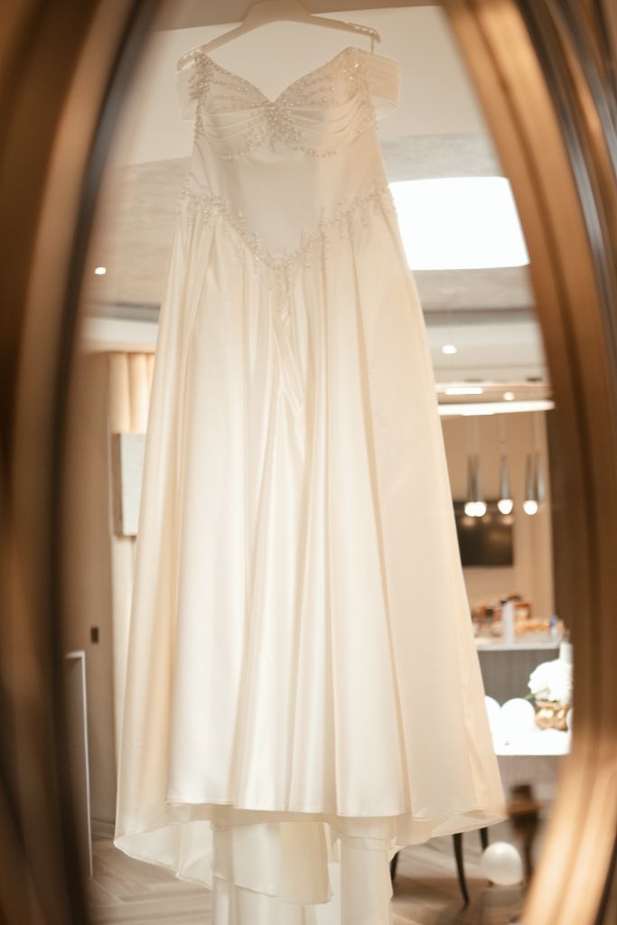 Elegant strapless white satin wedding gown with beaded bodice reflected in ornate gold mirror.