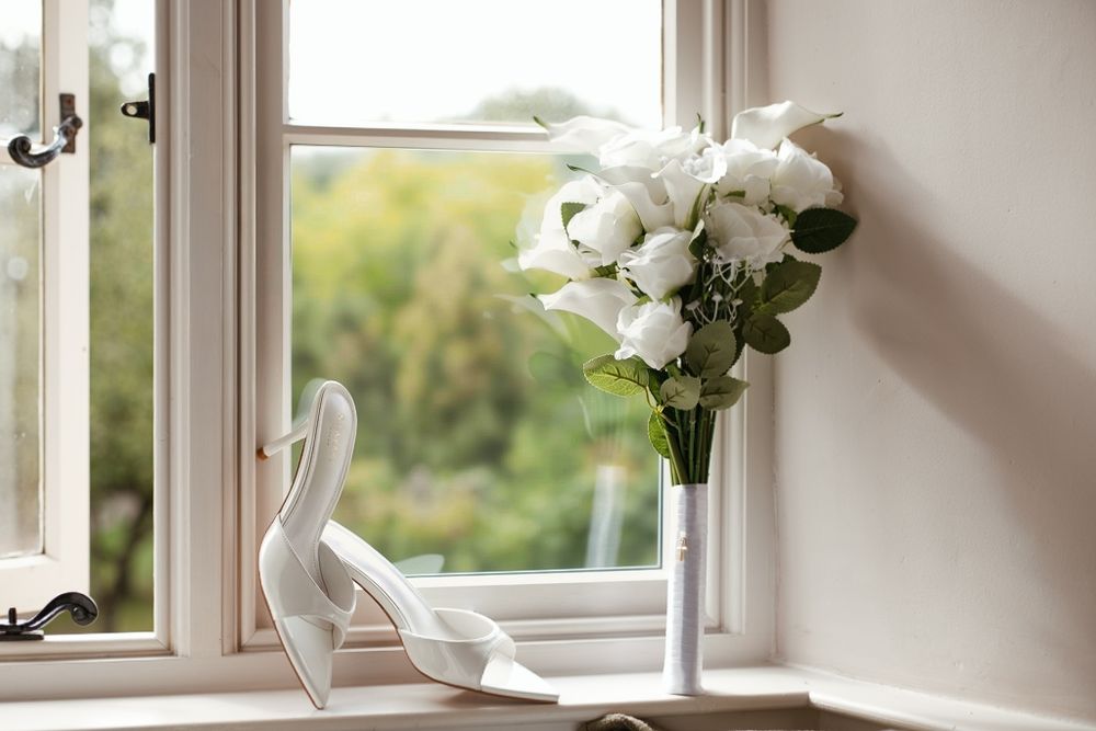 White bridal heels and a bouquet of white roses in a vase on a windowsill with a garden view.