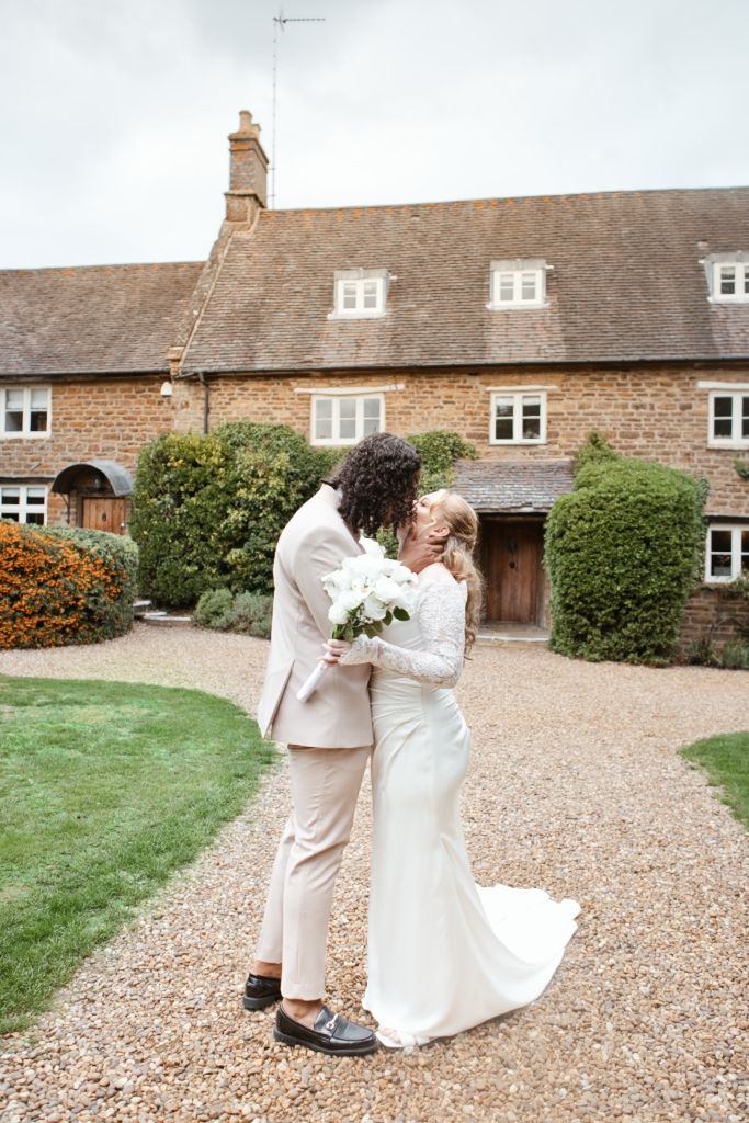 Bride and groom share a romantic kiss outside a rustic stone country house, bride holding white bouquet.