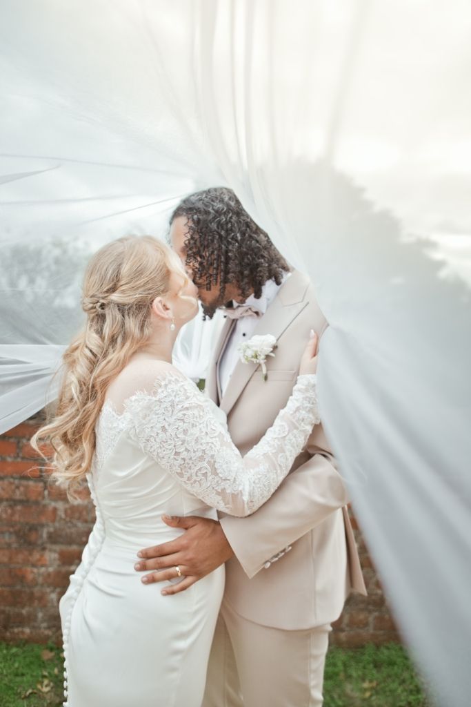Bride and groom share romantic moment under flowing white veil, interracial couple embracing in wedding attire outdoors.