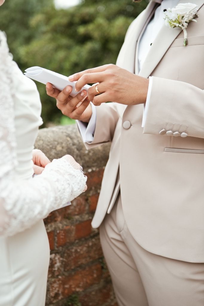 Groom in beige suit reading wedding vows from a card while bride in white lace dress listens outdoors.