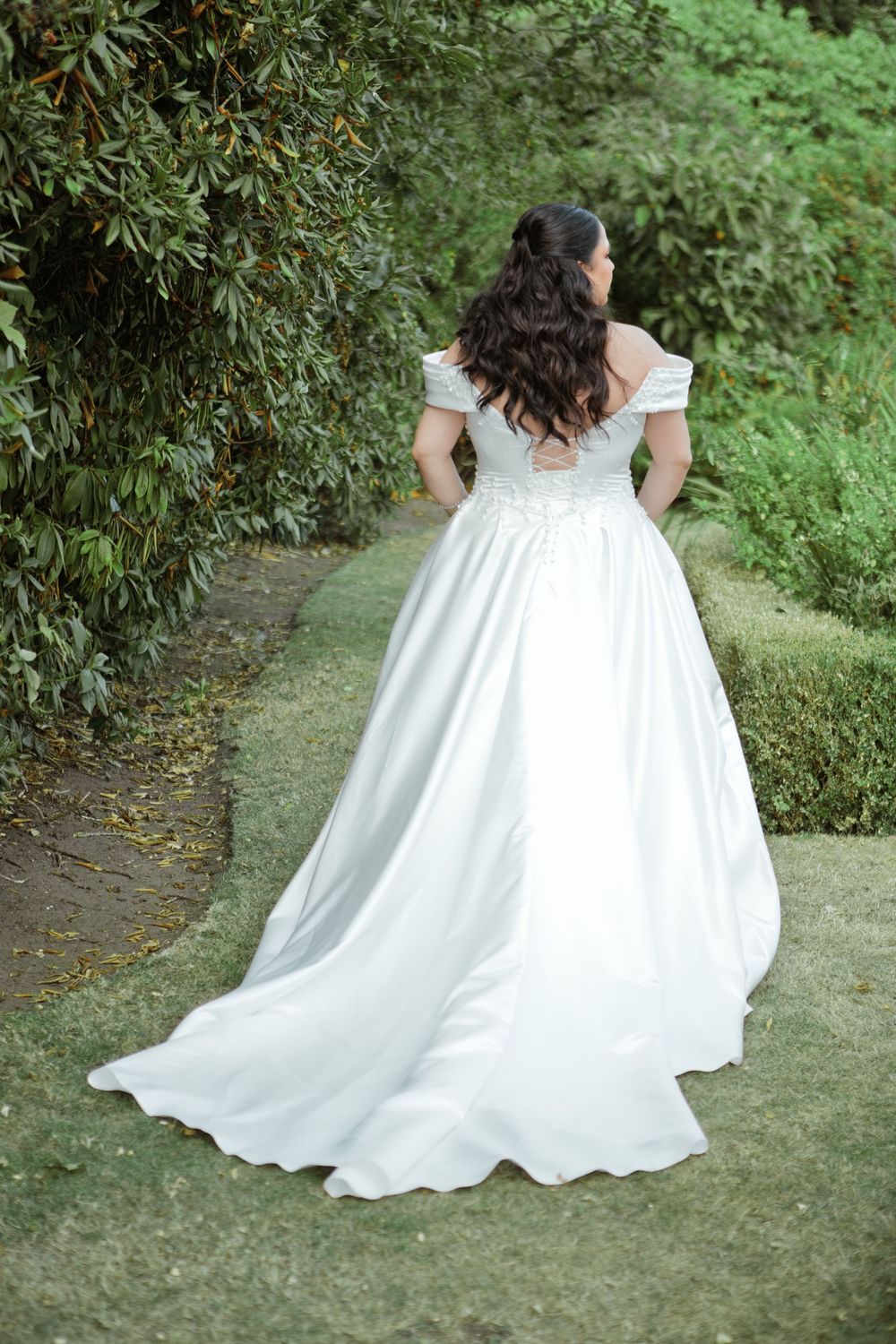 Bride in elegant off-shoulder white ball gown with train walking through lush garden greenery.