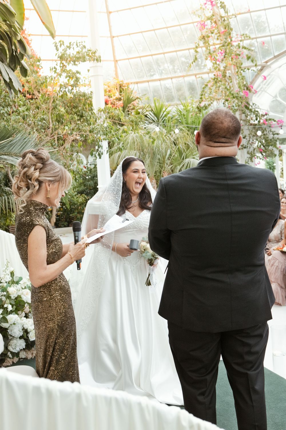 Bride laughing joyfully during heartfelt indoor greenhouse wedding ceremony with officiant and groom.