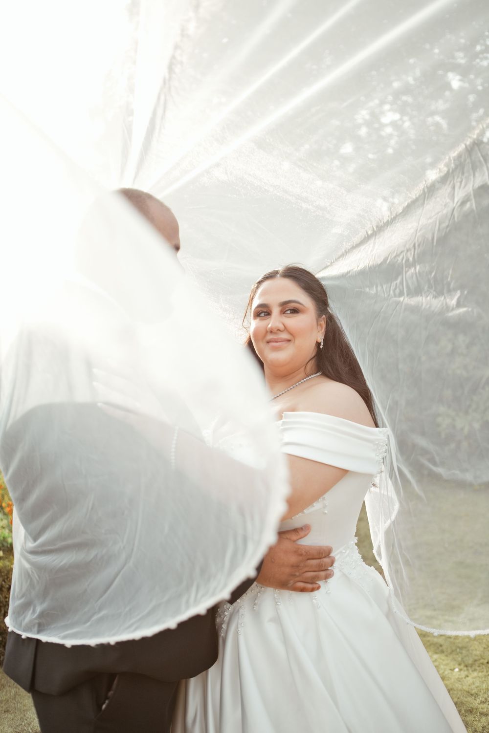 Bride in white off-shoulder gown smiles as groom lifts her veil outdoors in bright sunlight.