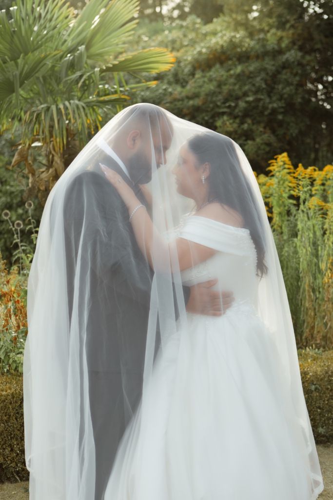 Bride and groom kissing under flowing veil in lush garden wedding setting.