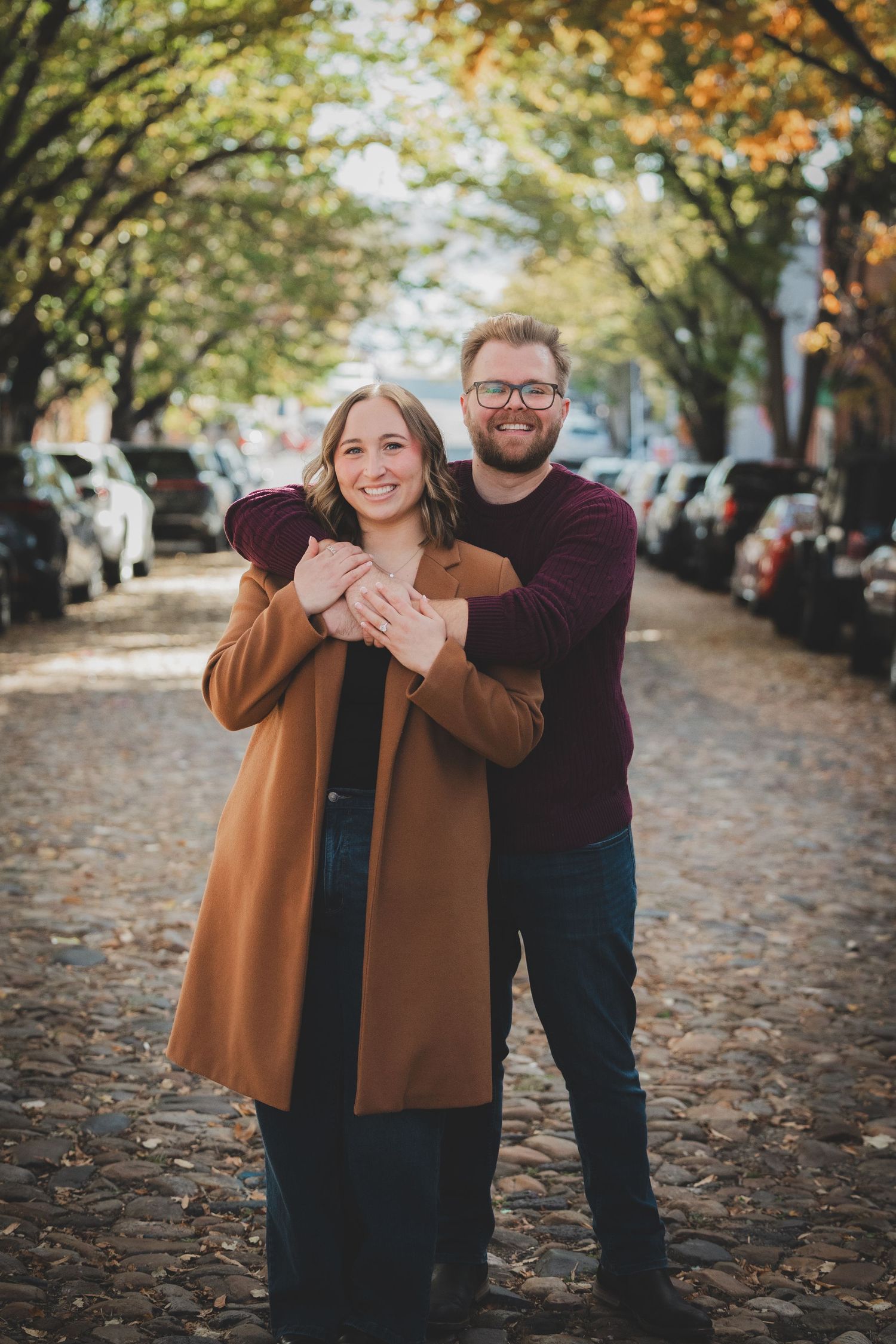 Newly engaged couple posing together on a tree-lined cobblestone street with golden fall foliage in Alexandria Virginia