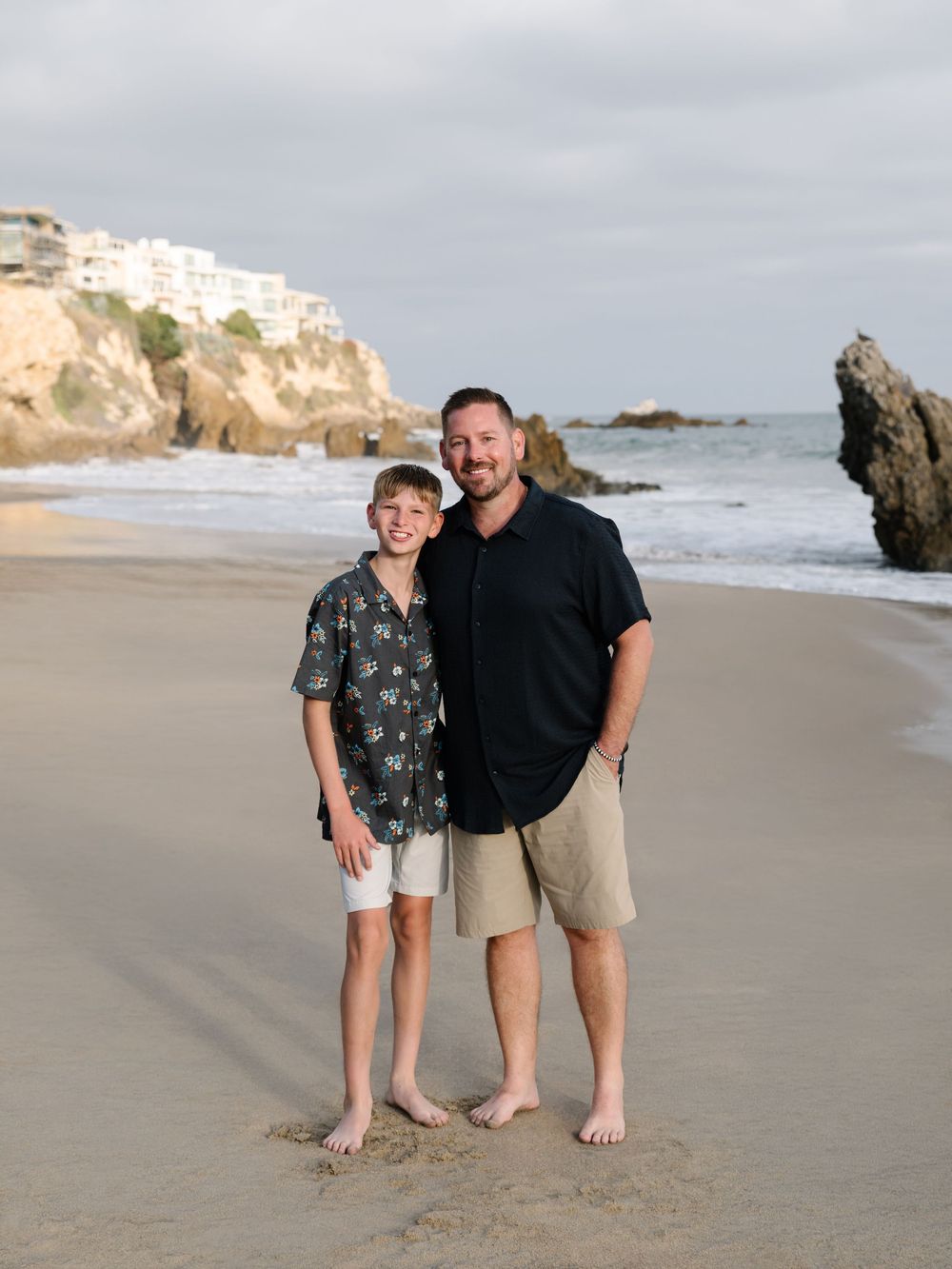 Father and son standing together on the beach with Newport Beach cliffs and ocean in the background.
