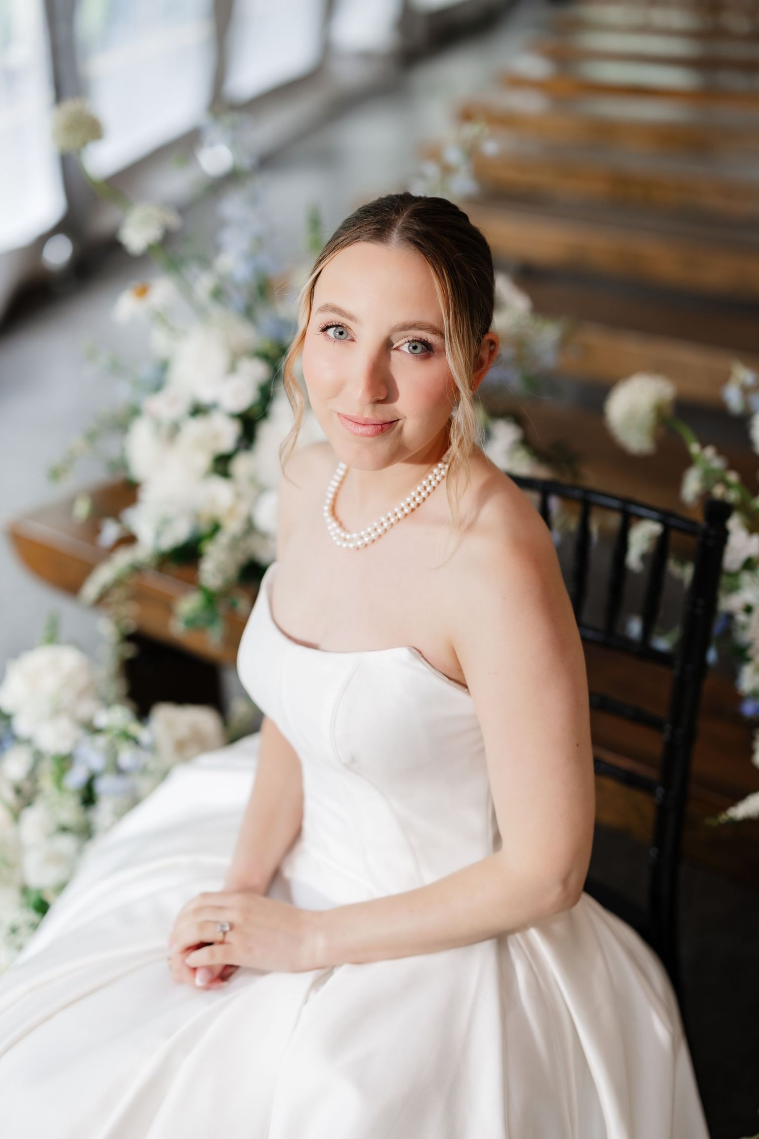 Beautiful bride in strapless white satin gown with pearl necklace seated among white and blue floral arrangements.