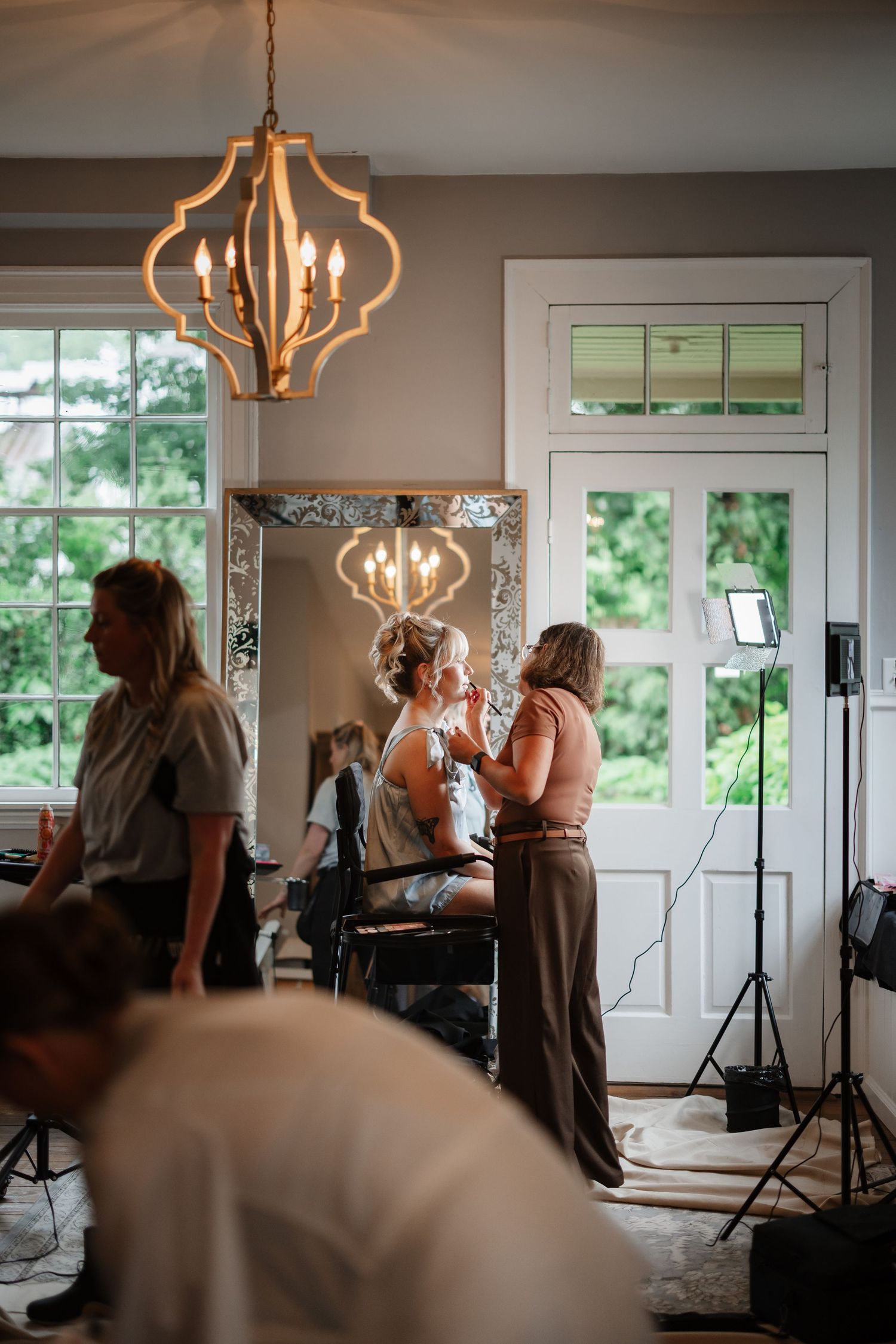 A bride gets her makeup done beneath a chandelier in a bright room with large windows and mirror.