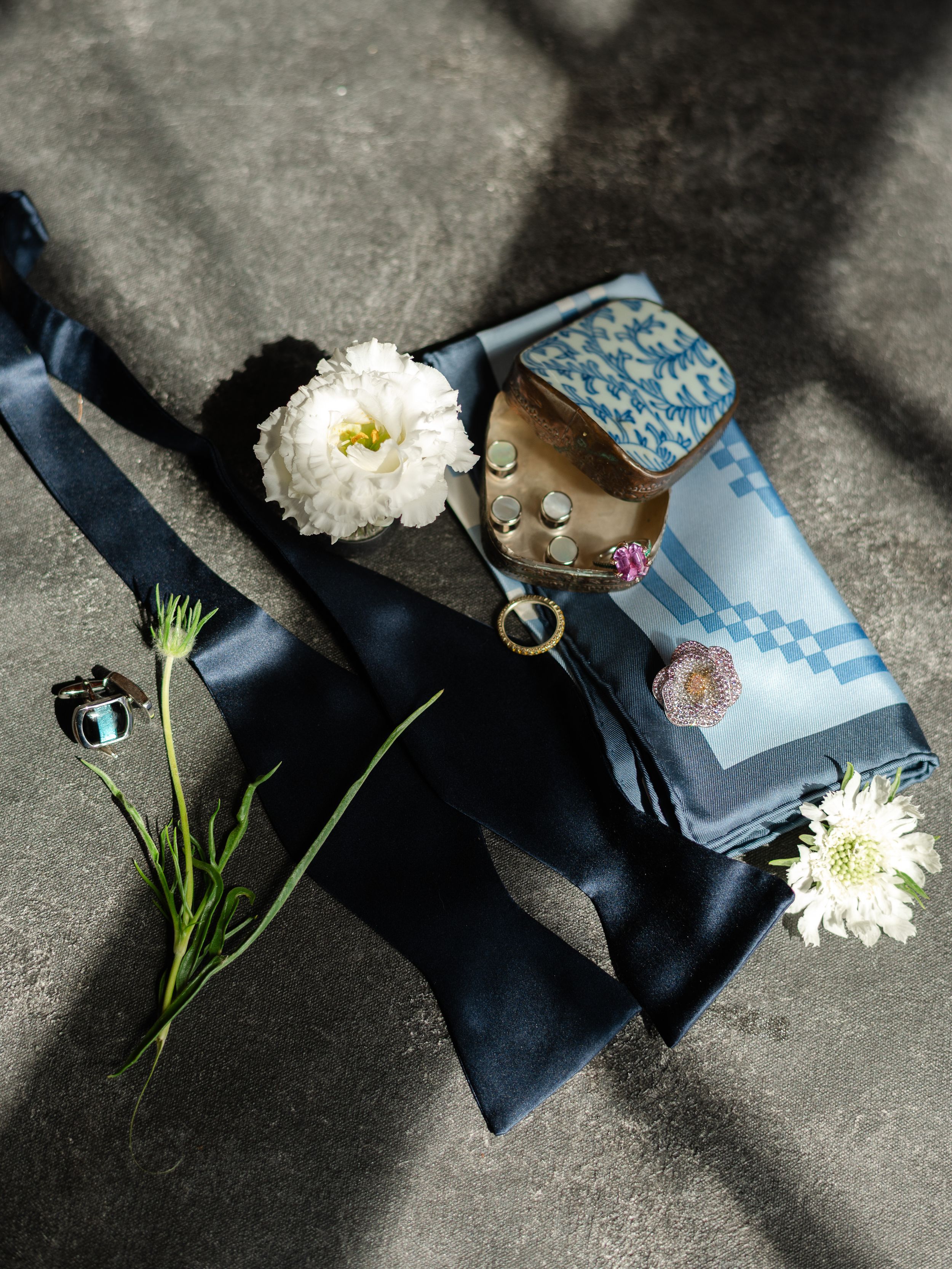 Flat lay of navy bow tie, blue patterned pocket square, floral brooch, and white flowers on gray surface.