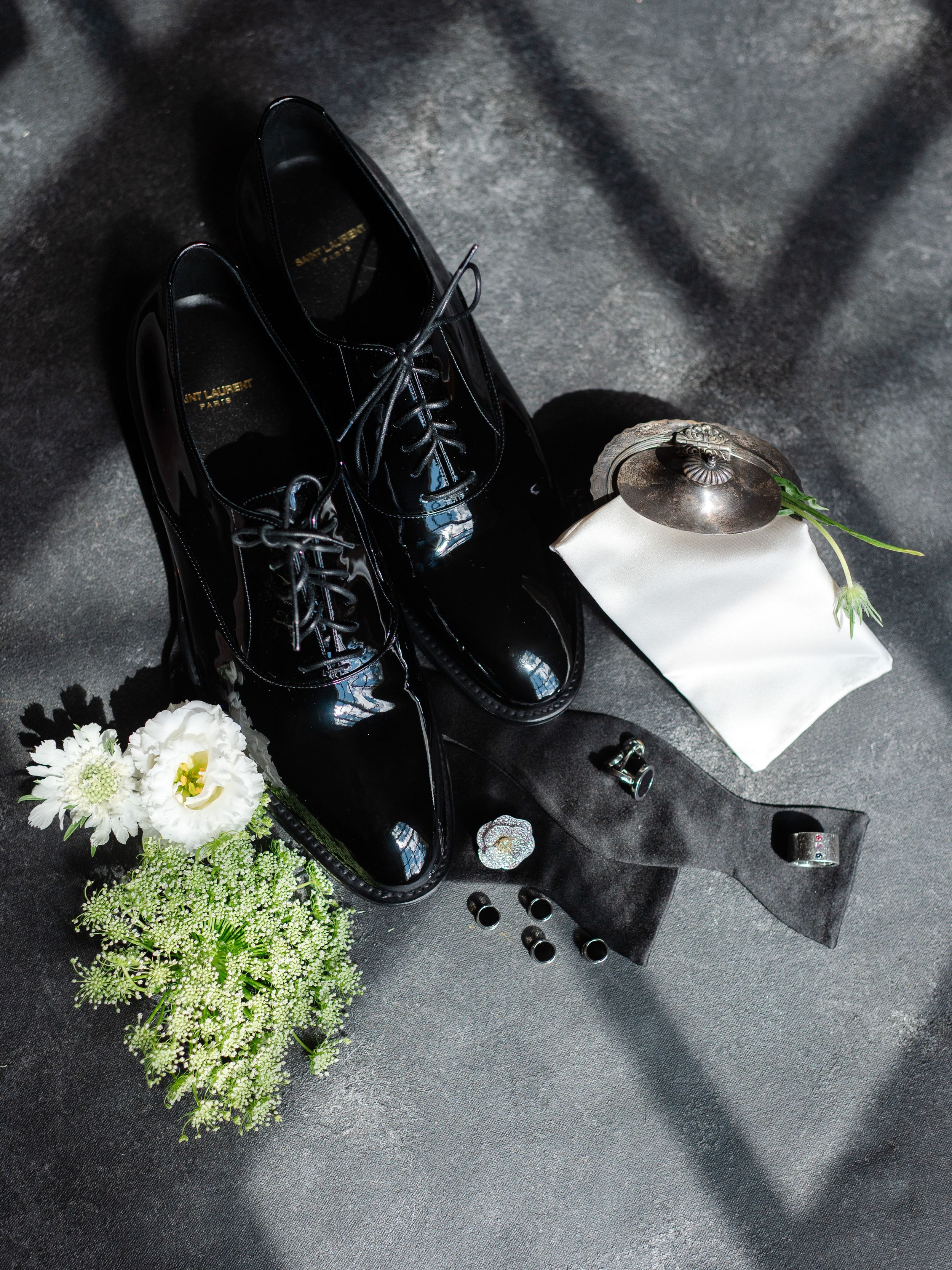 Black patent leather dress shoes with blue laces, white flowers, cufflinks, and a gray sock on a gray surface.